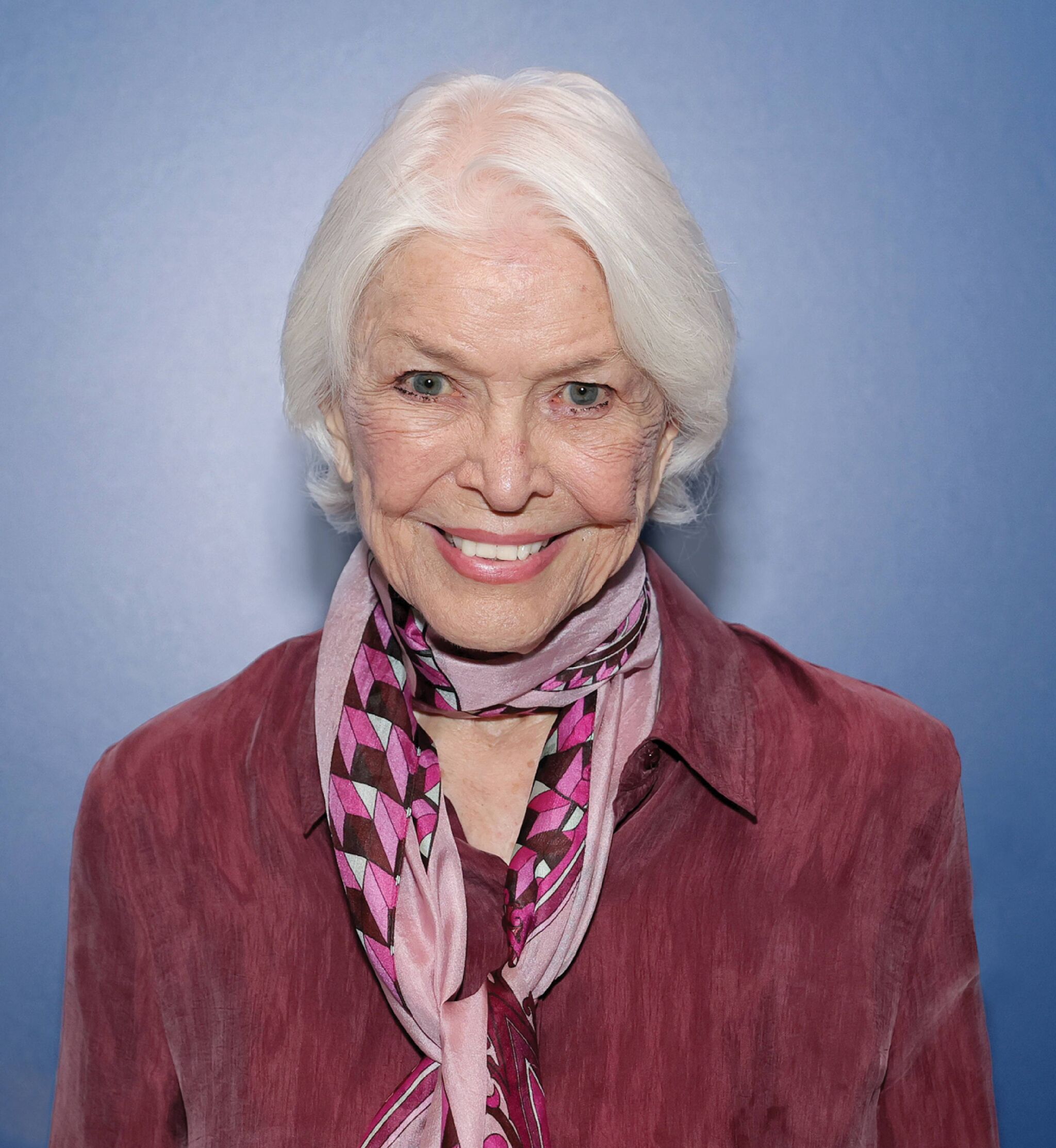 Portrait of Ellen Burstyn. She is smiling at the camera and wearing a maroon shirt with a pink and purple scarf wrapped around her neck