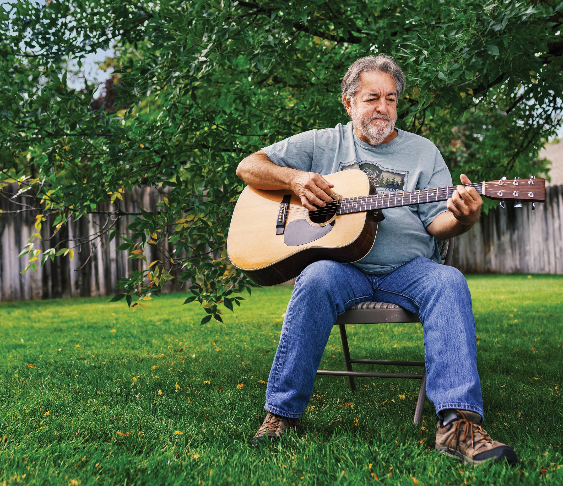 Photograph of Rubin Lopez Jr. sitting on a chair in a yard and playing the guitar.