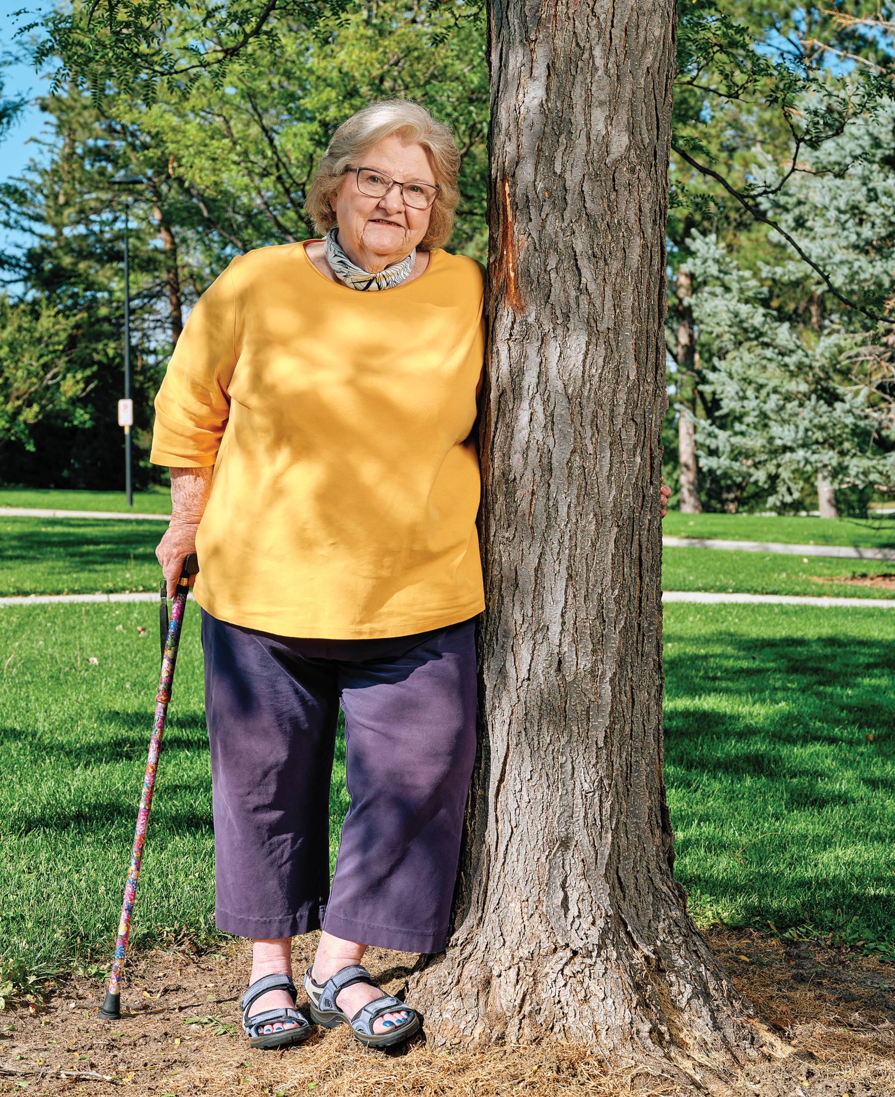 Photograph of Vicki Ivey in a yellow sweater and purple pants leaning against a tree in a garden.