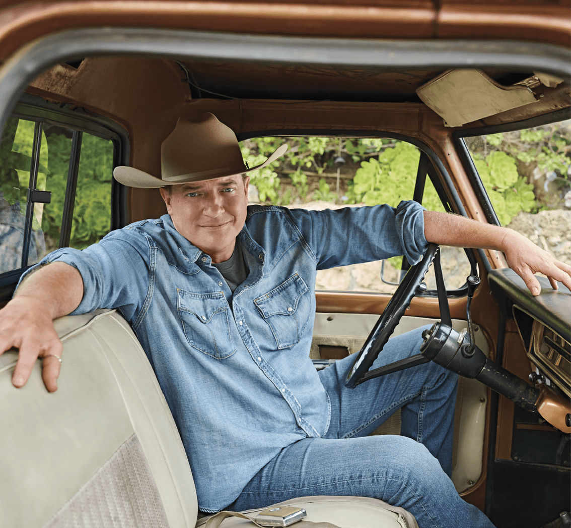 Image of Brendan Fraser in a denim button down, jeans and a cowboy hat sitting in a truck&nbsp;