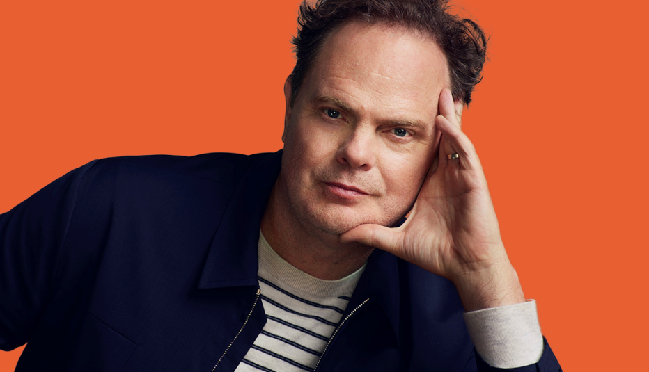 rainn wilson with face leaning against left hand; orange background