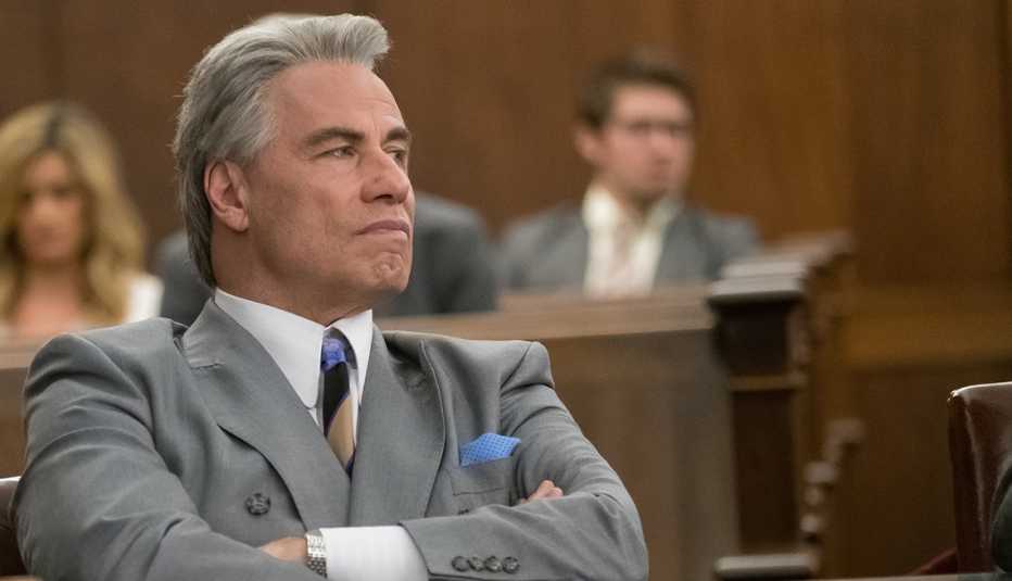 John Travolta sitting at a table with arms crossed.