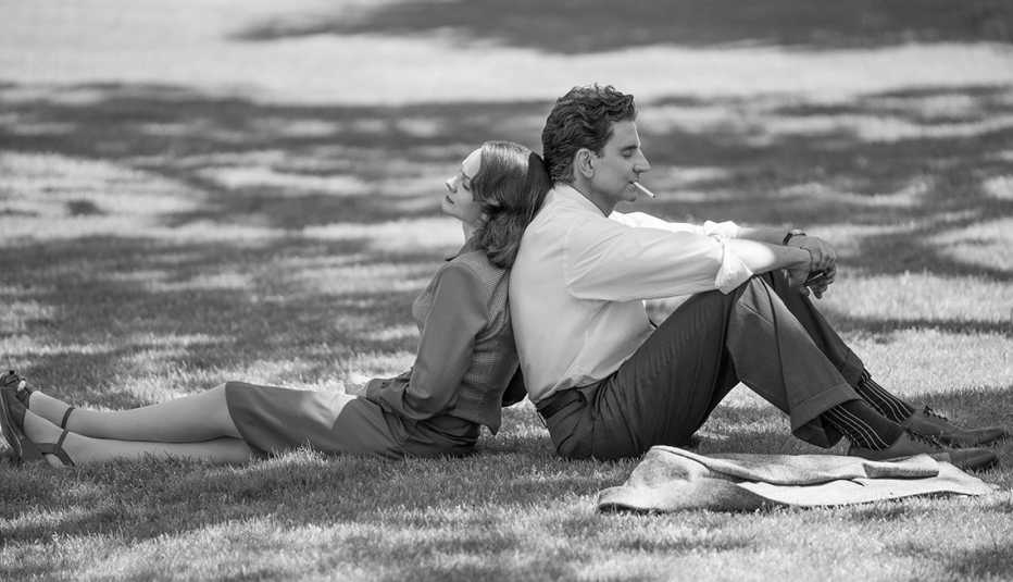 Carey Mulligan and Bradley Cooper sitting back-to-back on each other on the grass in a scene from the film "Maestro."