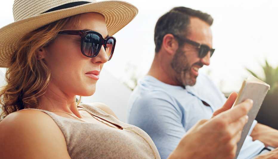 A couple wearing sunglasses sitting side by side on deck chairs and doing some reading outdoors