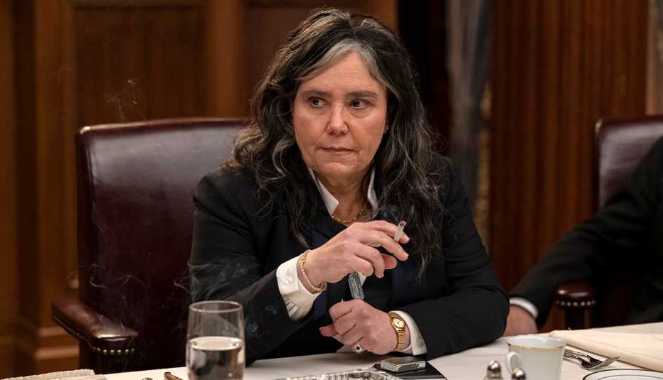 alex borstein sitting in a chair at a table with a cigarette in her hand in the marvelous mrs maisel