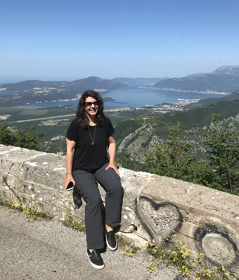 Elaine Glusac sits on a wall in front of a scenic overlook of mountains and water
