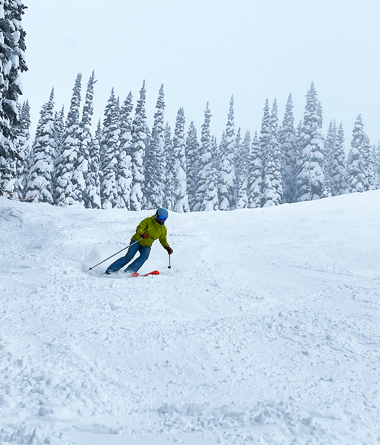 skier in chartreuse jacket and blue helmet skiing down a snowy mountain with snow-covered trees behind her