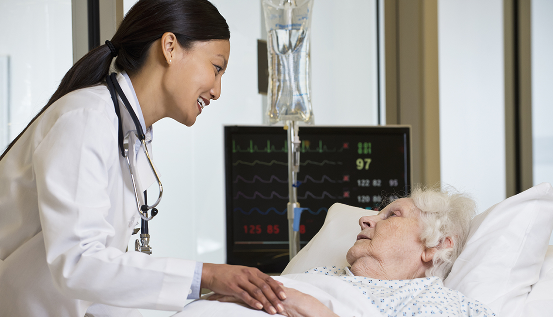 Female doctor comforting older woman lying in a hospital bed. Heart monitor and saline drip in the background. 