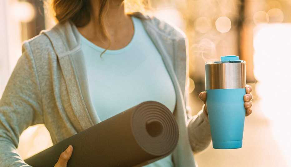 A woman holding a yoga mat in one hand and a coffee mug in the other