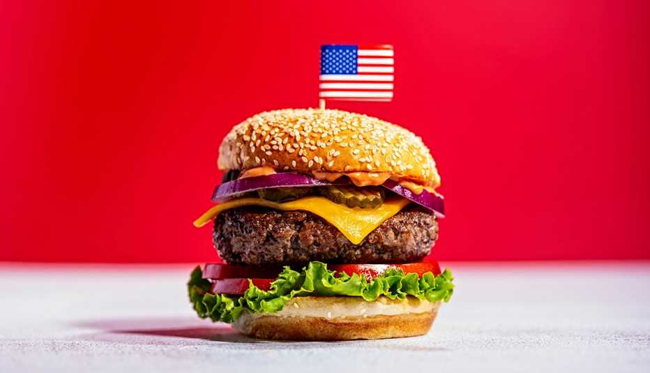 A hamburger with meat, cheese, vegetables on a sesame seed bun with an american flag toothpick stuck in the top