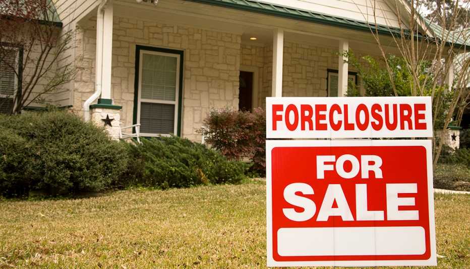 White stone home with green roof and shrubbery in front.  A red and white sign in the foreground reads "Foreclosure - For Sale."
