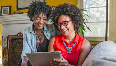 two women sitting on a couch looking at a tablet