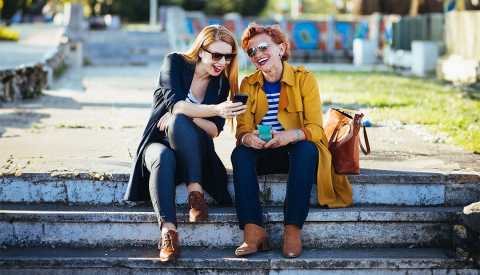 two women sitting on a step outside looking at a mobile phone and laughing