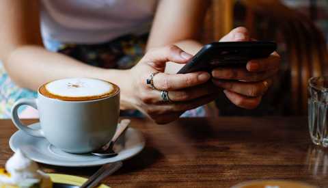 woman's hands holding a smart phone with a cub of coffee next to them