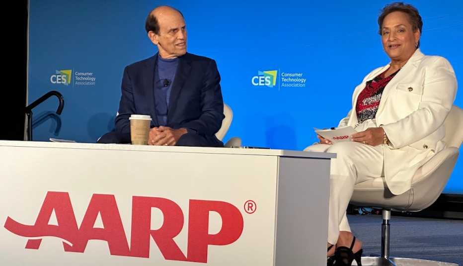 michael milken and aarp ceo jo ann jenkins sit behind a desk that reads aarp