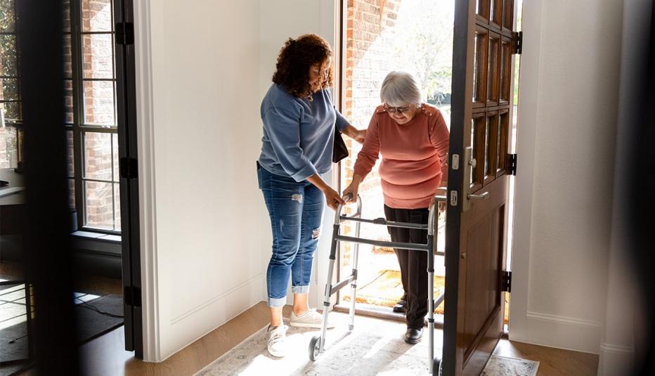 A healthcare worker helps an elderly woman walk using a rollator. Indoors, home setting