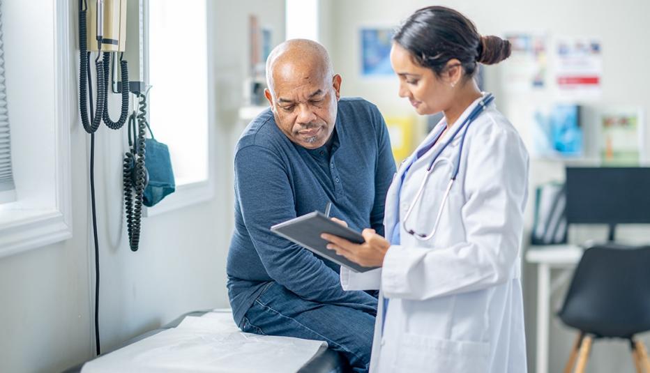 A senior gentleman of African decent sits up on an exam table during a routine check-up. His female doctor is holding out a tablet as they review some recent test results together.