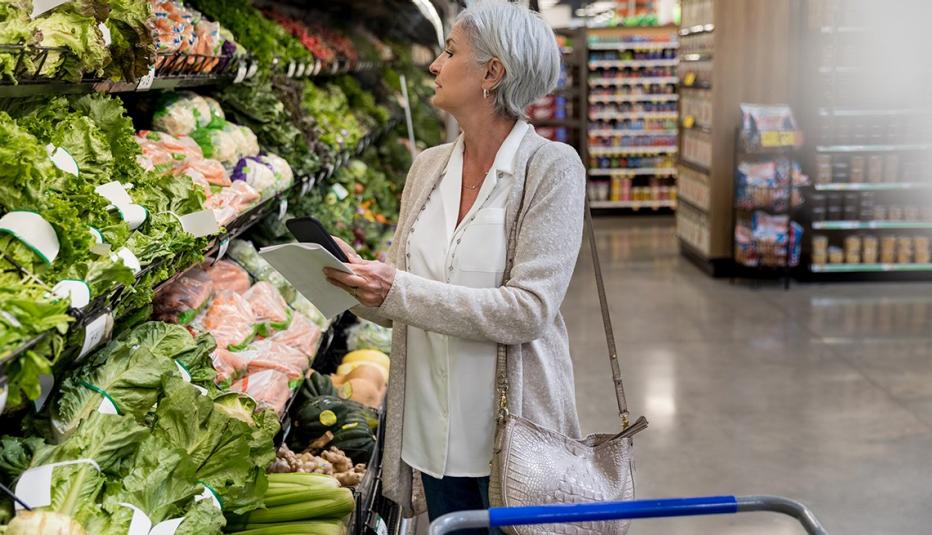 Mature woman holds smartphone while shopping for leafy greens in local supermarket.