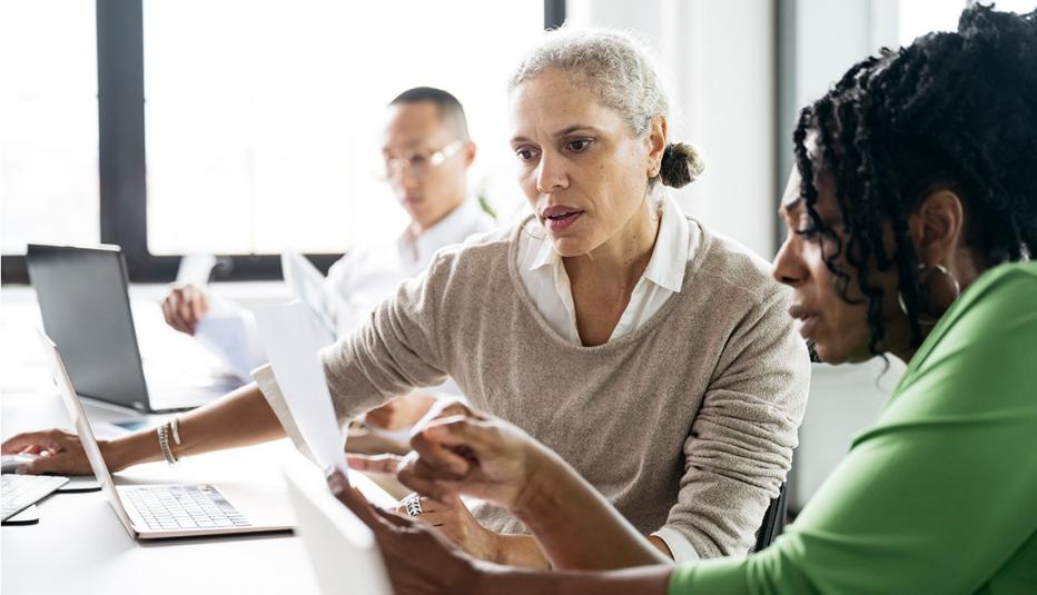 Two mature business women sitting at desk using laptop and discussing important document. Female office workers studying paperwork in office, collaboration, strategy, teamwork