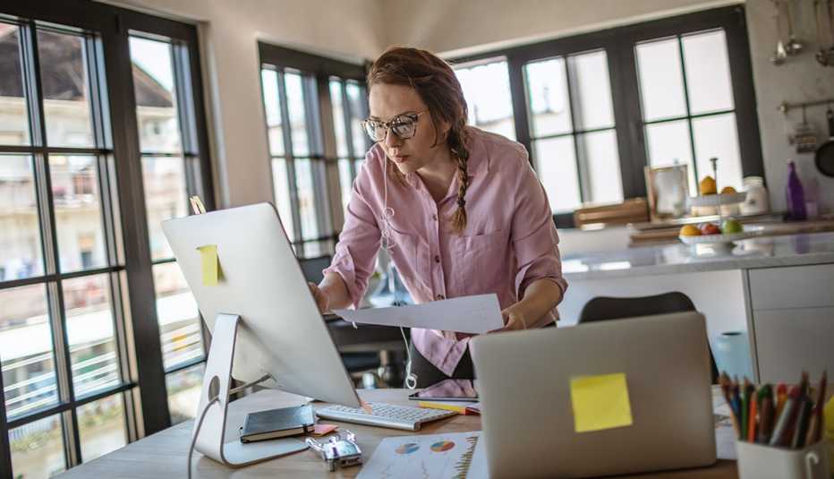 Mujer con un papel en la mano y viendo su computadora en la cocina de su casa.