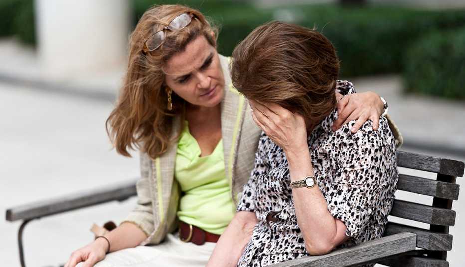 Mujer consuela a su madre colocándole una mano en su hombro. Ambas están sentadas en la banca de un parque