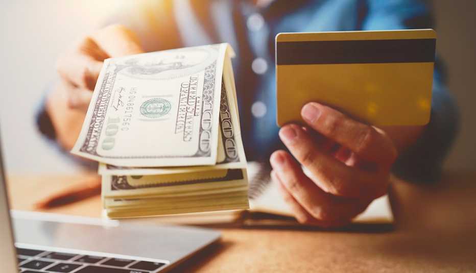Manos frente a una computadora sostiene billetes de 100 dólares y una tarjeta de crédito.