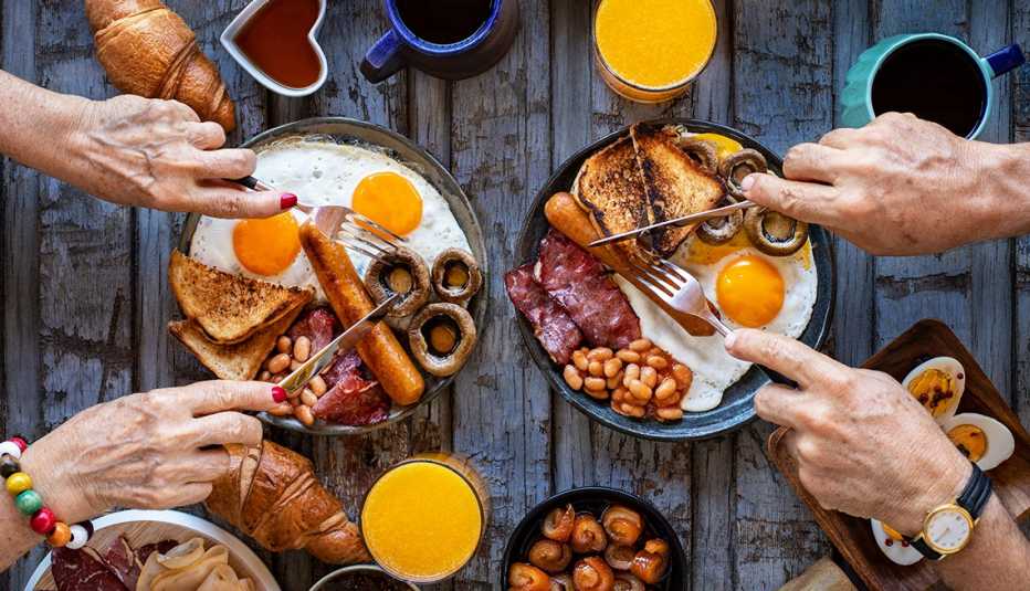 Mesa servida con platos de un desayuno tradicional y manos de personas con cubiertos sobre ellos.