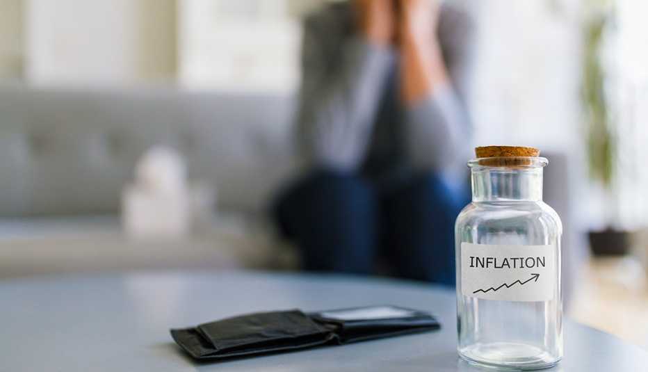 Mujer sentada en un sofá se cubre las manos con la cara y frente a ella una mesa con un frasco vacío que dice inflación