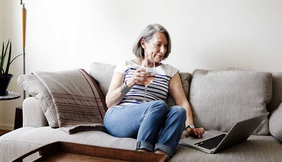 Mujer mayor sentada en una sofa con una copa en la mano y viendo un computador portátil