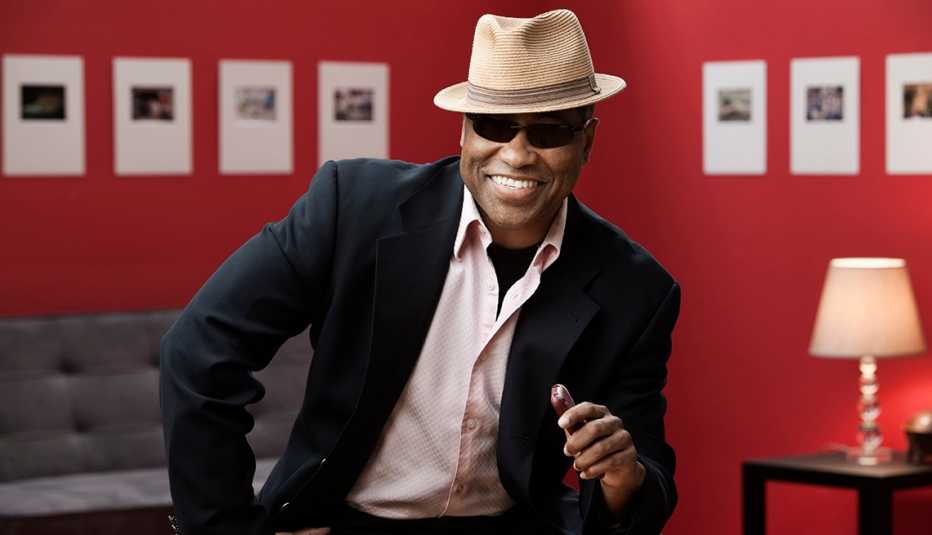 Un hombre elegante y sonriente llevando un sombrero, anteojos, traje, chaqueta y camisa con cuello.