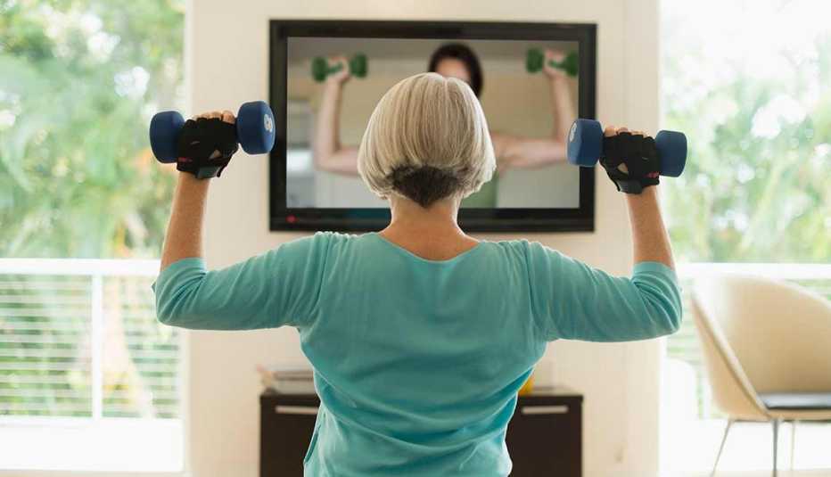 Mujer ejercitándose con dos mancuernas frente a un televisor