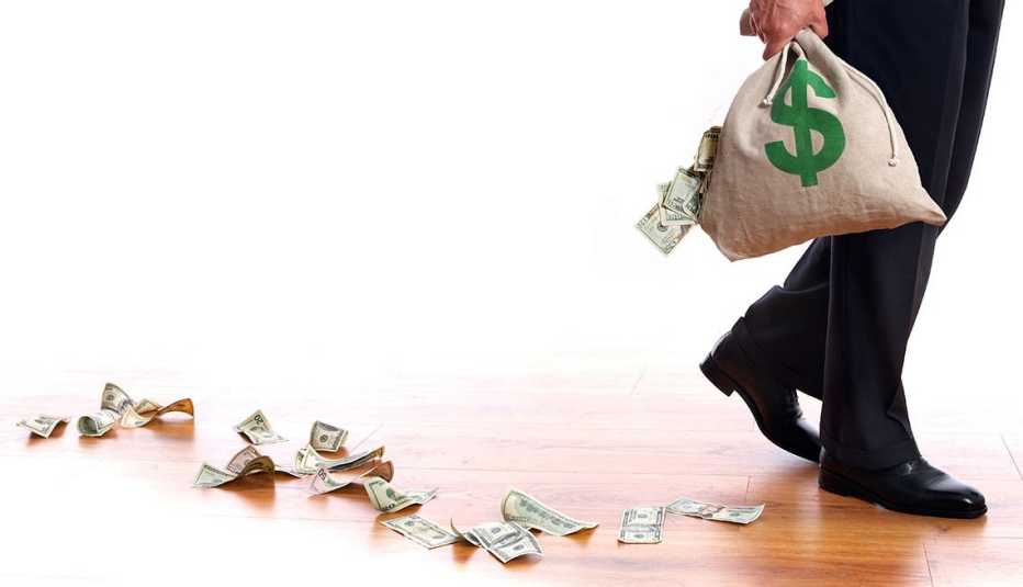 Pies de un hombre y una mano que sostiene una bolsa con dinero que se va regando en un piso de madera