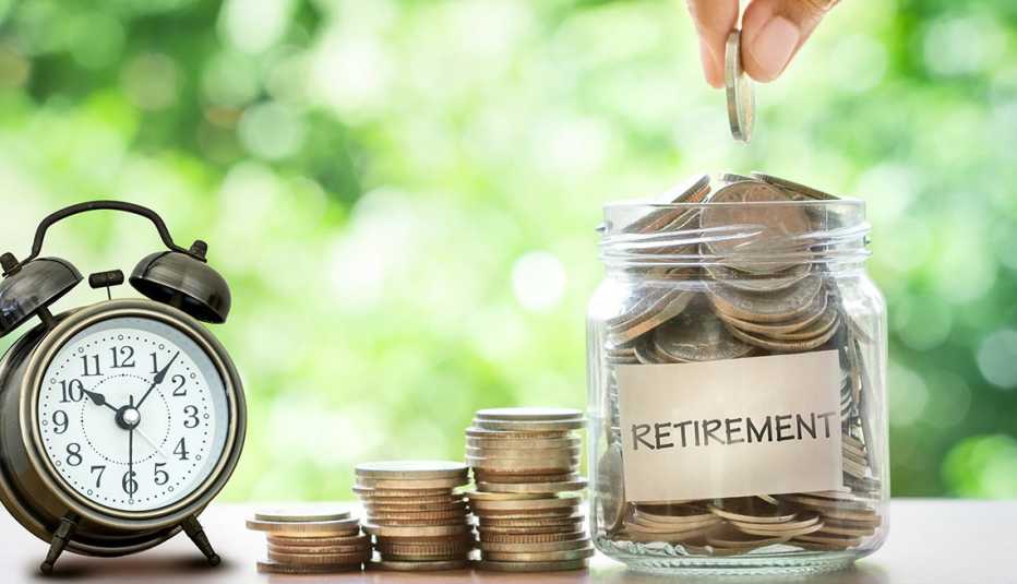 Mano colocando dinero en una jarra que dice jubilación al lado de un reloj despertador