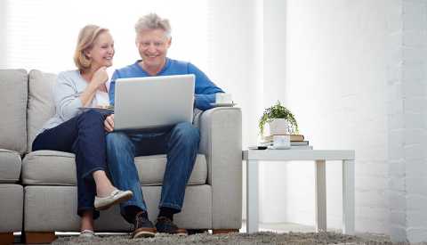 Pareja mayor sentada en sofa, sonriendo enfrente se un computador portátil