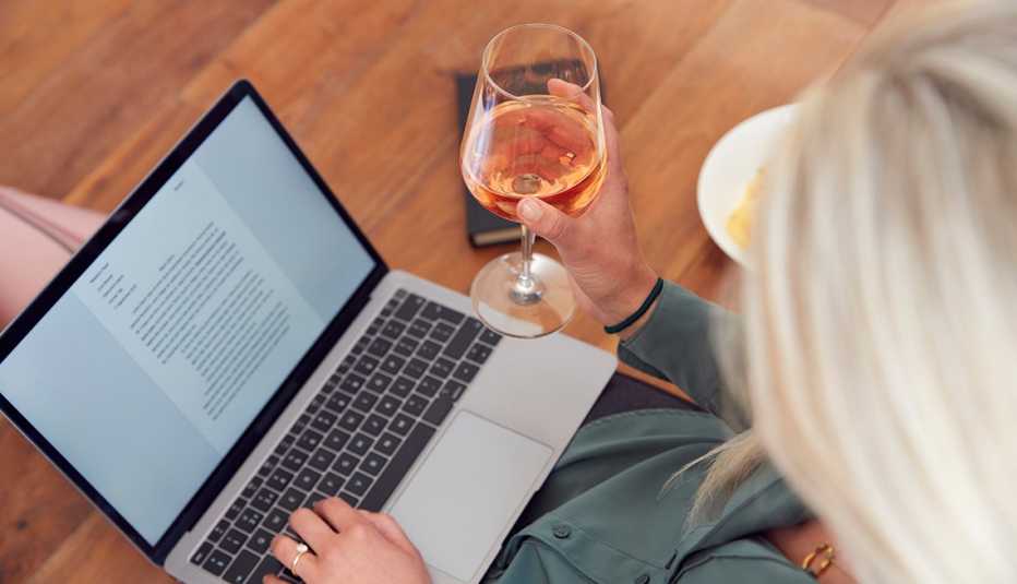 Una mujer trabaja desde su casa en una computadora, mientras sostiene una copa de vino