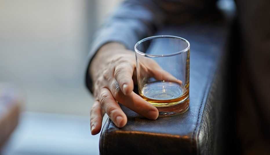 Mano de un hombre en el descanso de una butaca sostiene un vaso con licor