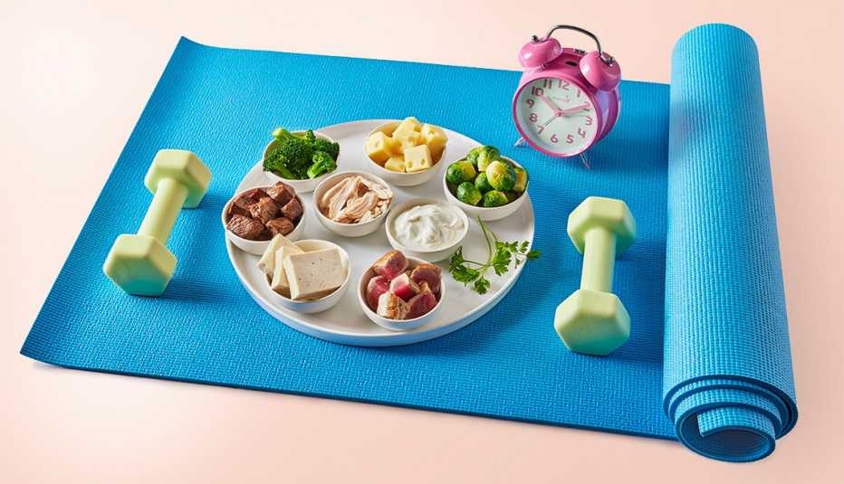 Una estera de yoga y sobre ella hay un reloj, varios alimentos nutritivos y un  par de mancuernas