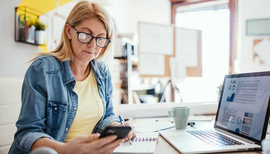 Mujer mira su teléfono mientras está sentada frente a la computadora en su casa.