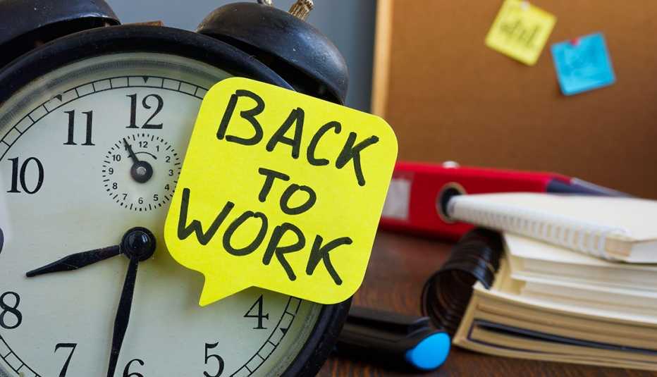 Reloj despertador al lado de una pizarra de chorcho, libretas, y encima un letrero que dice regreso al trabajo
