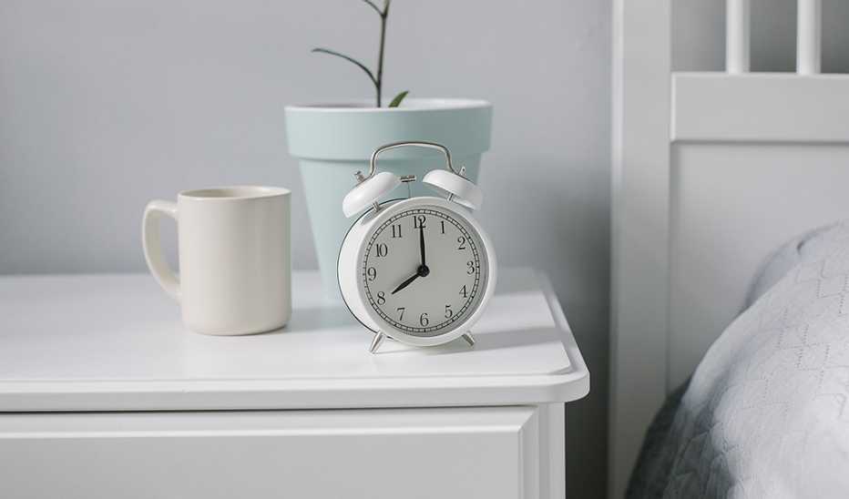 A white alarm clock on a nightstand by a bed